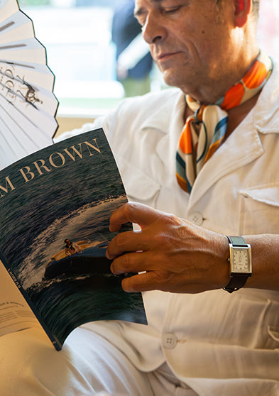 TWG069000: Un hombre que lleva un reloj WM Brown x Timex mientras lee la revista WM Brown y sostiene un abanico plegable abierto.