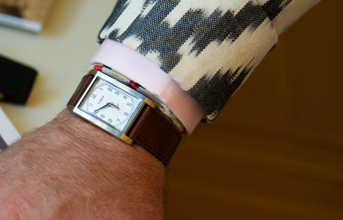 Reloj rectangular TWG069000 WM Brown x Timex, con esfera blanca y correa de piel marrón, que se lleva junto con un puño rosa y una chaqueta estampada.