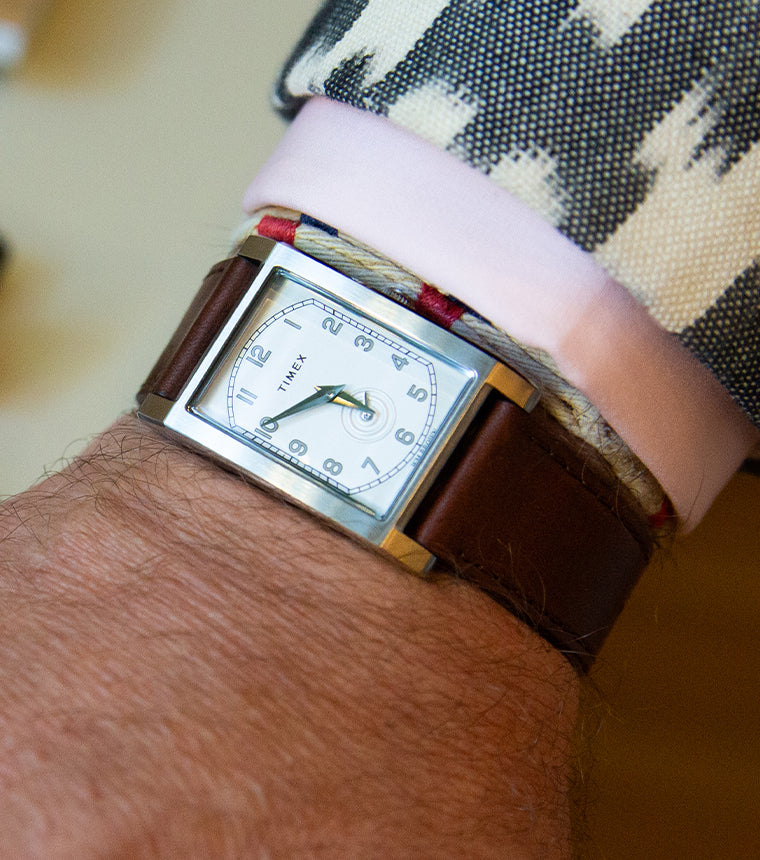 Reloj rectangular TWG069000 WM Brown x Timex, con esfera blanca y correa de piel marrón, que se lleva junto con un puño rosa y una chaqueta estampada.