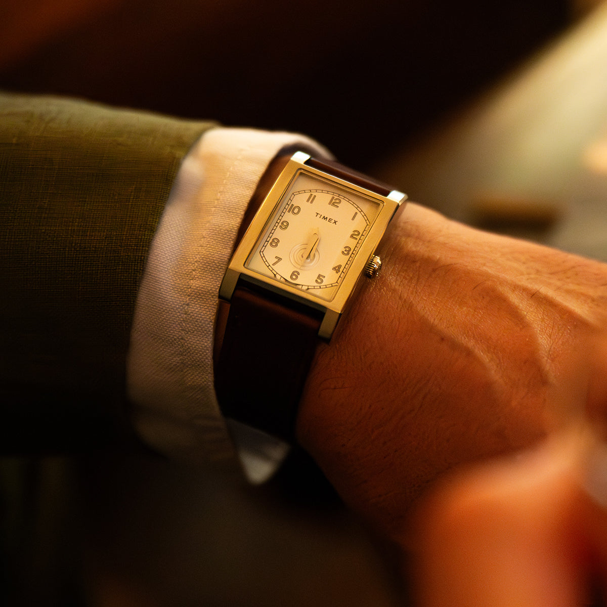 TWG069000 Primer plano de un reloj WM Brown x Timex con caja rectangular y correa de piel marrón, colocado junto al puño de una camisa bajo una luz cálida.