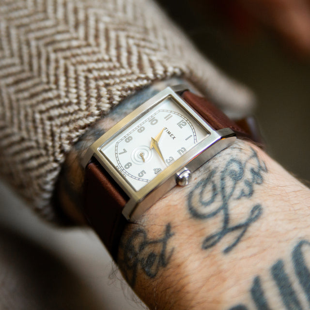 Reloj TWG069000 WM Brown x Timex con esfera blanca, caja rectangular en tono plateado y correa de piel marrón en una muñeca tatuada.
