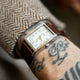 Reloj TWG069000 WM Brown x Timex con esfera blanca, caja rectangular en tono plateado y correa de piel marrón en una muñeca tatuada.