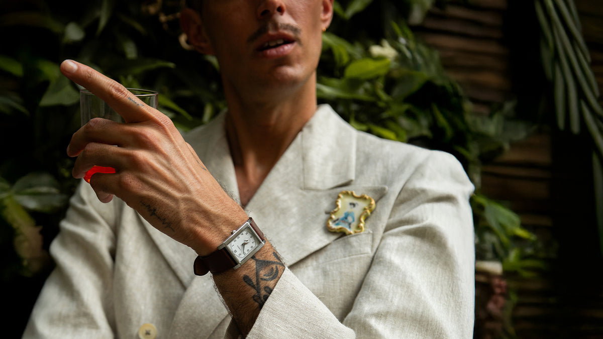 Modelo TWG069000 con un traje color crema, que lleva un reloj WM Brown x Timex con correa de piel marrón y sostiene una copa.