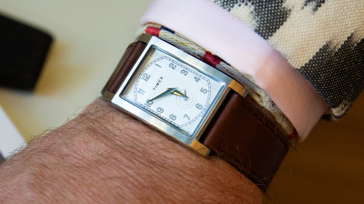 TWG069000 Primer plano de un reloj rectangular WM Brown x Timex con correa de piel marrón, que se lleva debajo de un puño rosa y una chaqueta estampada.