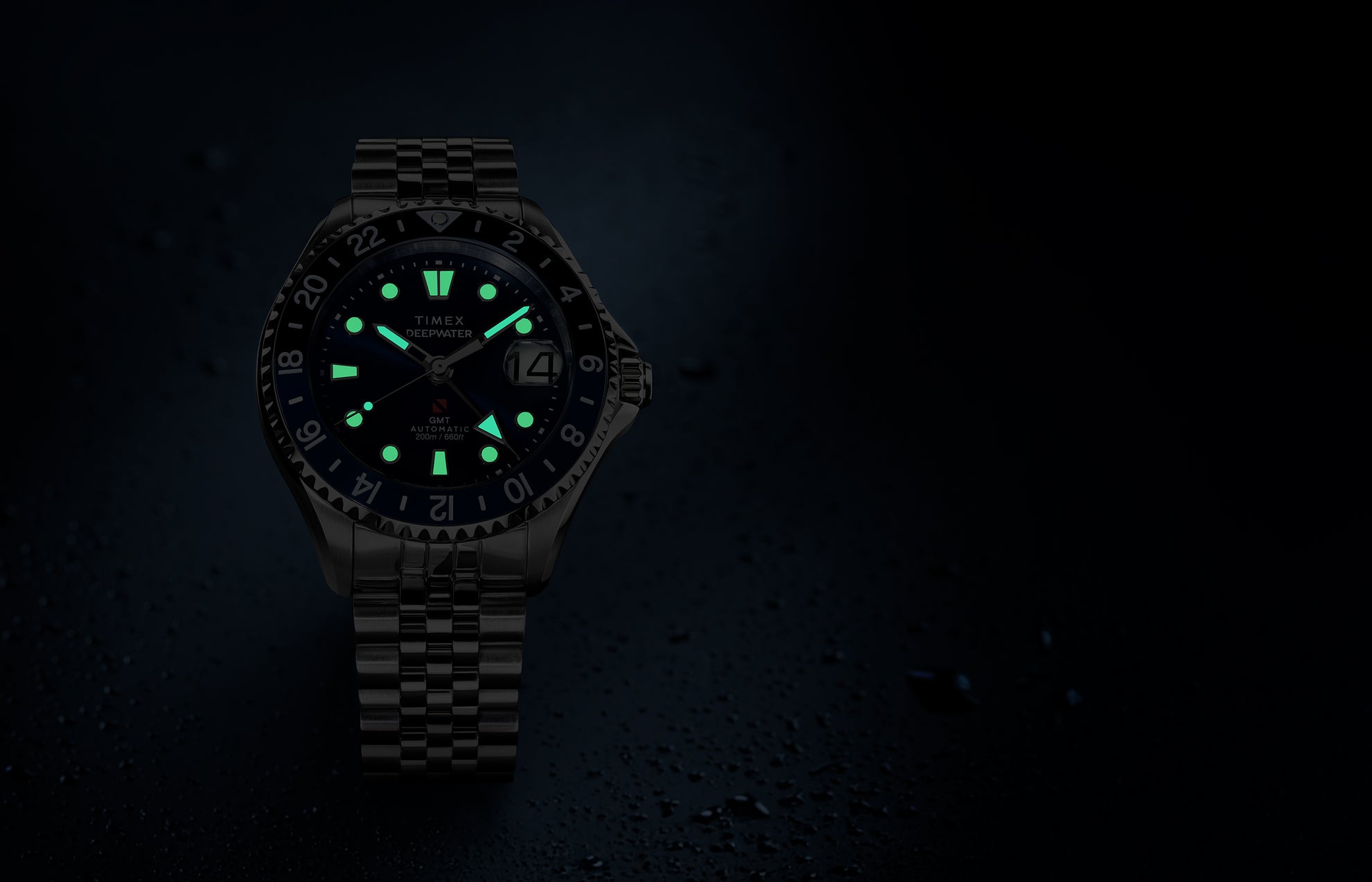 Timex Deepwater Meridian 200 Automatic GMT watch with a blue dial and glowing luminous hands and hour markers, displayed against a dark, water-droplet background.