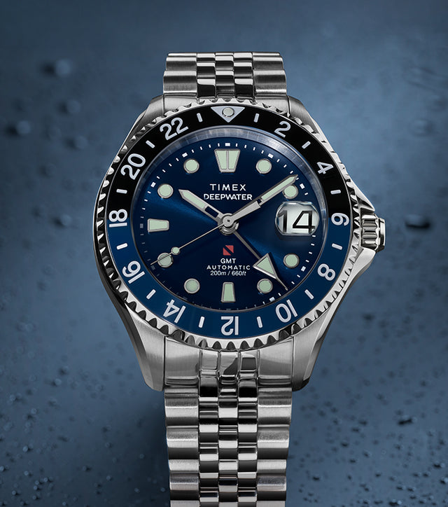 Timex Deepwater Meridian 200 Automatic GMT stainless steel watch featuring a blue sunray dial, GMT bezel, magnified date window, and jubilee-style bracelet on a textured blue background.