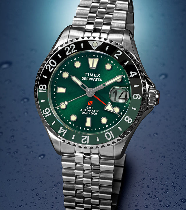 Timex Deepwater Meridian 200 Automatic GMT stainless steel watch featuring a green sunray dial, black-and-green GMT bezel, magnified date window, and jubilee-style bracelet on a water-droplet background.