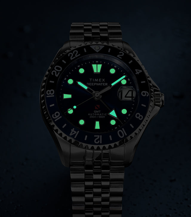 Timex Deepwater Meridian 200 Automatic GMT watch with a blue dial and glowing luminous hands and hour markers, displayed against a dark, water-droplet background.