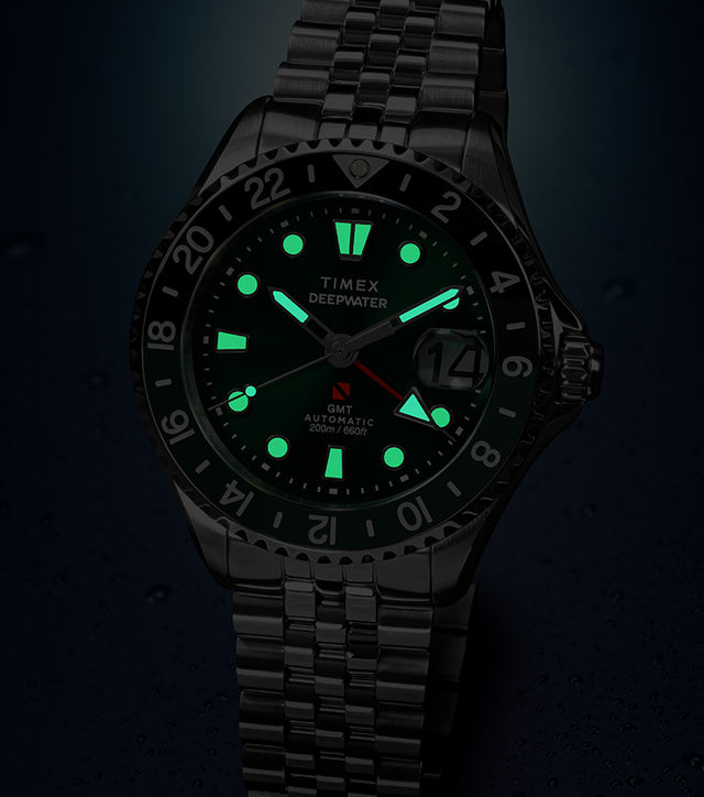 Timex Deepwater Meridian 200 Automatic GMT watch with a green dial and glowing luminous hands and hour markers, displayed against a dark blue background with water droplets.