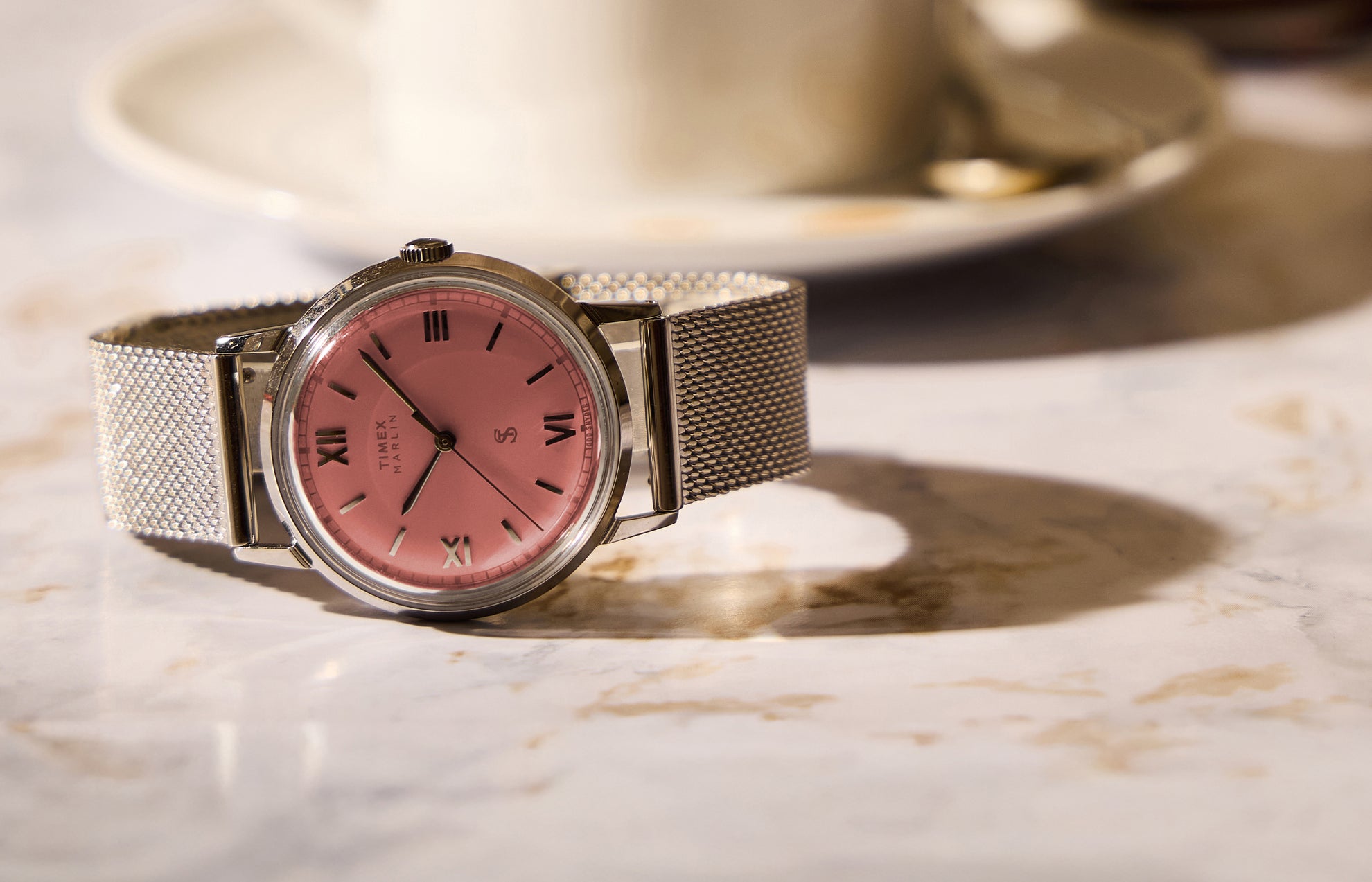 Todd Snyder x Timex Marlin® Mesh watch with a pink dial and stainless steel mesh bracelet displayed on a marble table beside a coffee cup.