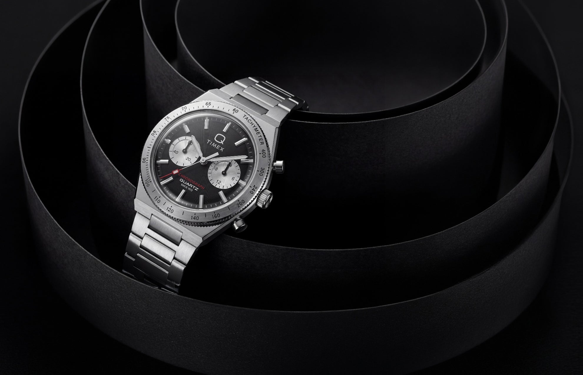 Q Timex Continental Chronograph with a black dial, silver-tone subdials, and stainless-steel bracelet displayed against sculptural black curves.