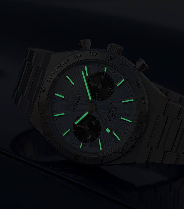 Q Timex Continental Chronograph in low light with green luminous hands and hour markers glowing against a dark blue background.