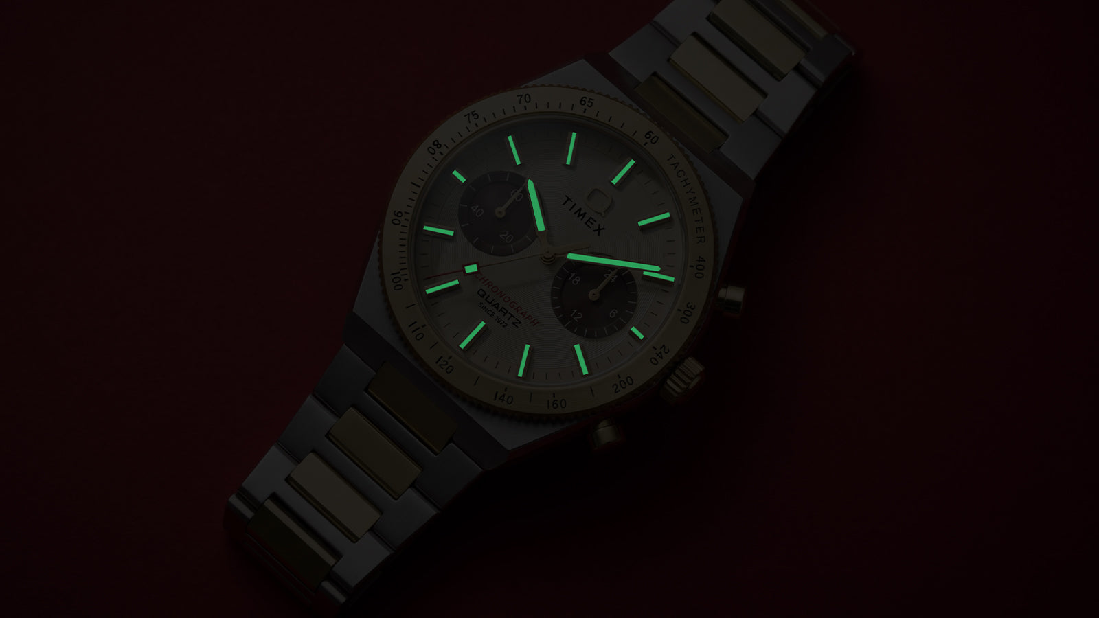 Q Timex Continental Chronograph in low light with green luminous hands and hour markers glowing against a dark red background.