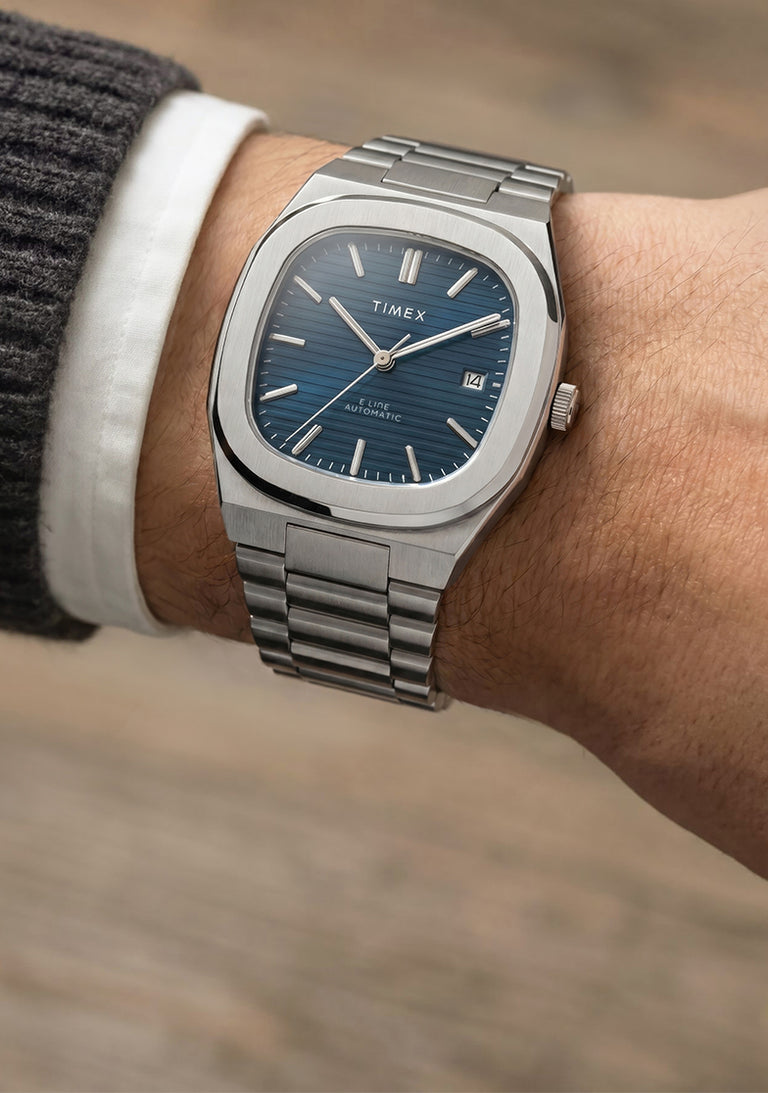 Primer plano del reloj Timex® Automatic E Line de 38 mm, de acero inoxidable, con esfera azul texturizada y ventana de fecha, puesto en la muñeca.