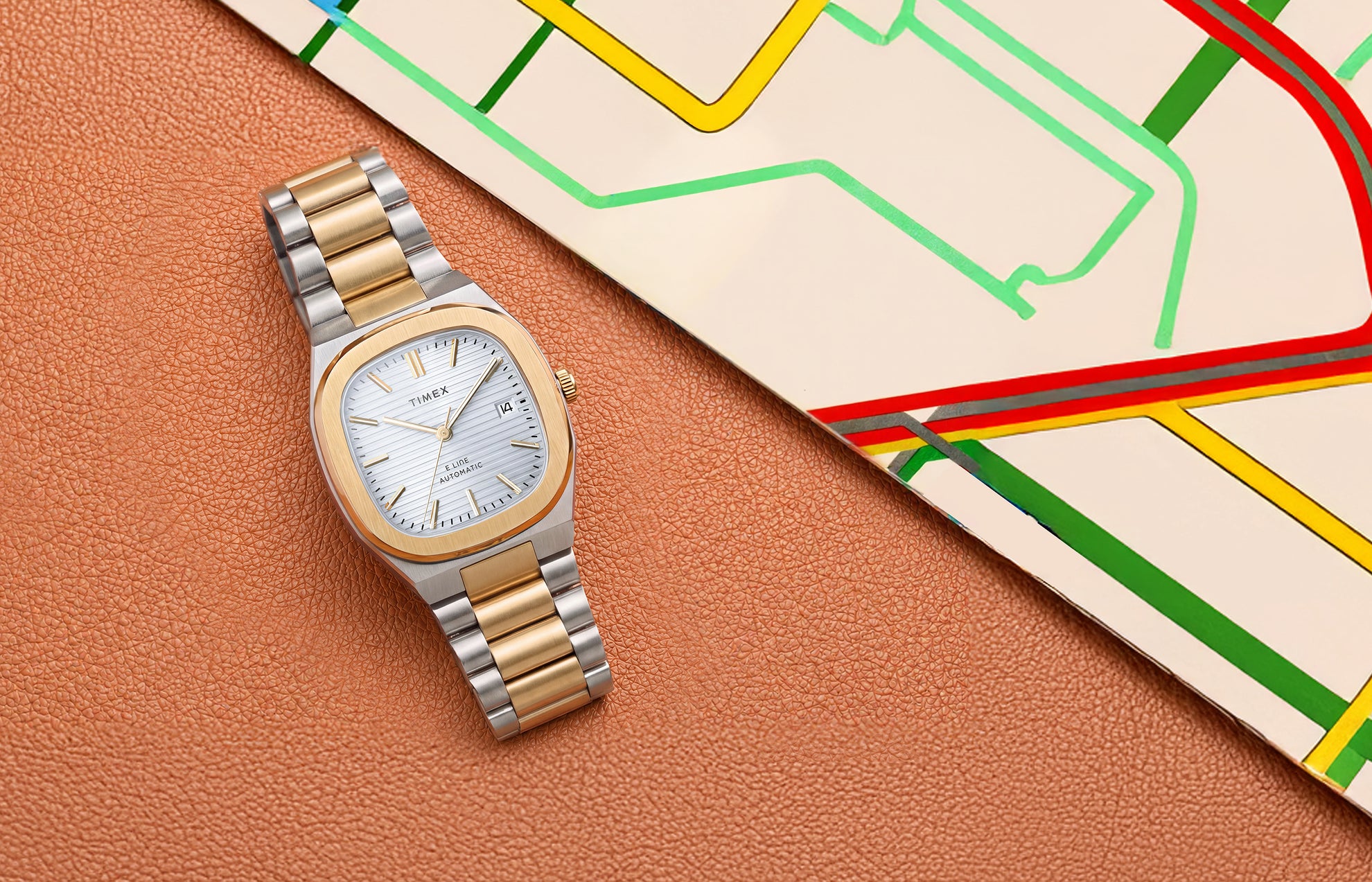 Timex® Automatic E Line watch with a silver dial and two-tone bracelet displayed on a textured brown surface beside a colorful transit map.