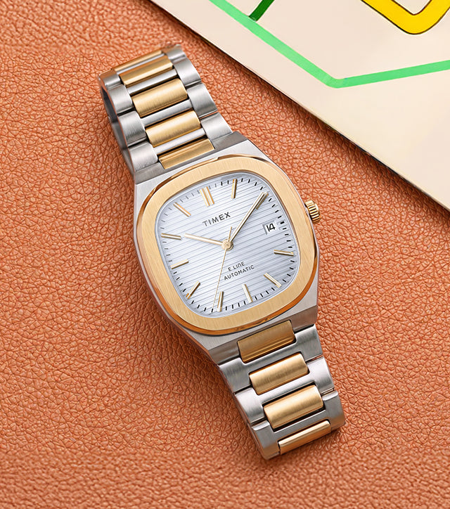 Timex® Automatic E Line 38mm watch with a two-tone stainless steel bracelet, gold-tone bezel, silver textured dial, and date window displayed beside a colorful transit map.