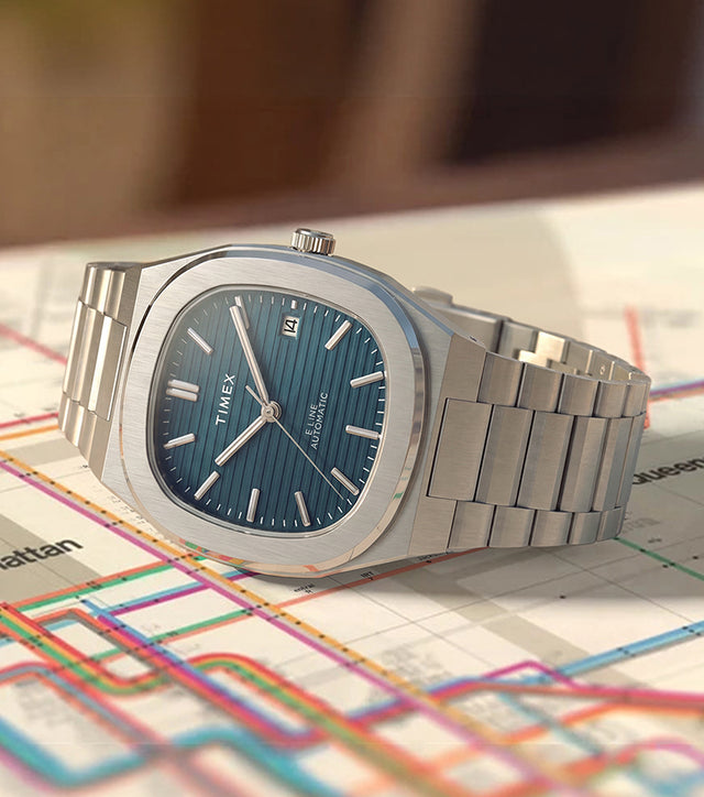 Reloj Timex® Automatic E Line de 38 mm con brazalete de acero inoxidable, esfera azul con textura y ventana de fecha representada en un mapa de transporte público.
