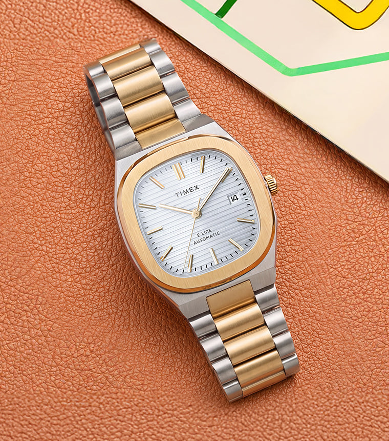 Timex® Automatic E Line watch with a silver dial and two-tone bracelet displayed on a textured brown surface beside a colorful transit map.
