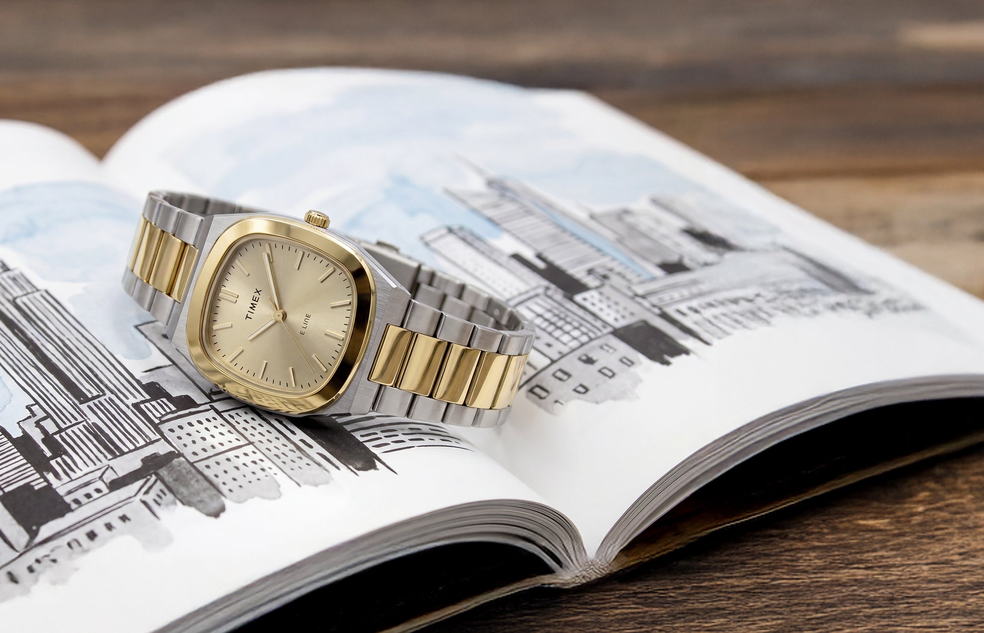 Timex® Quartz E Line 30mm watch with a two-tone stainless steel bracelet, gold-tone bezel, and champagne dial displayed on an open book with an illustrated cityscape.