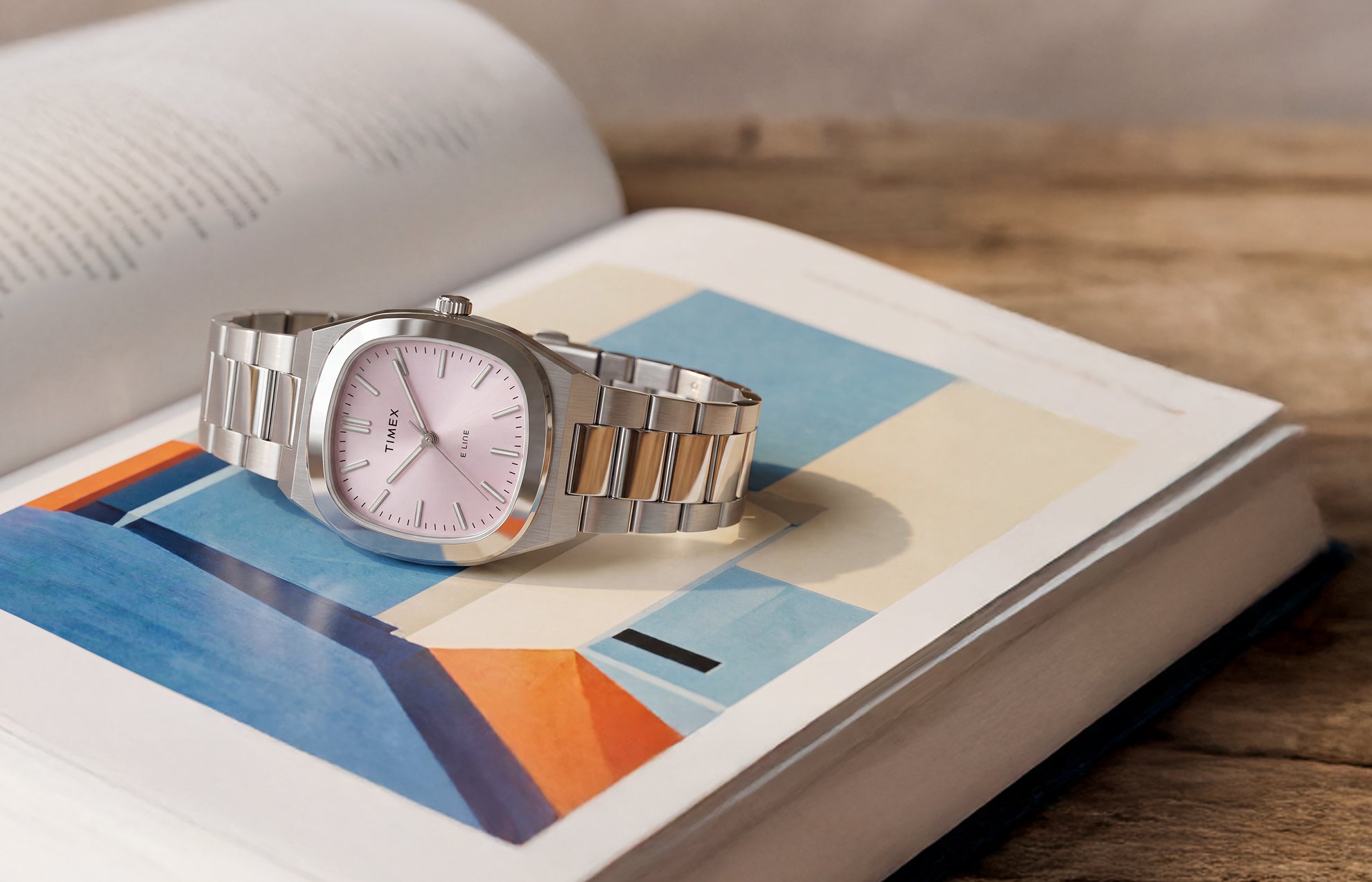 Reloj Timex® Quartz E Line de 30 mm con brazalete de acero inoxidable y esfera rosa con acabado rayos de sol, expuesto sobre un libro abierto con ilustraciones de colores.