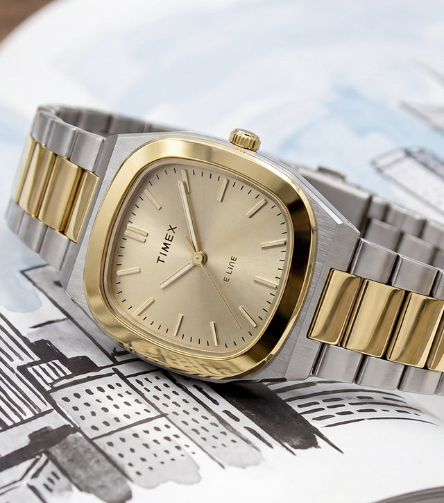 Timex® Quartz E Line 30mm watch with a two-tone stainless steel bracelet, gold-tone bezel, and champagne dial displayed on an open book with an illustrated cityscape.