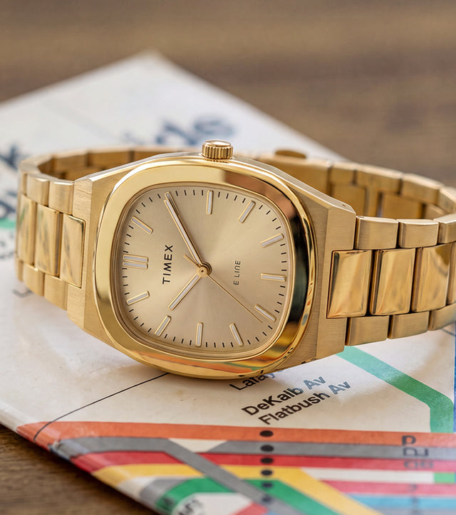 Reloj Timex® Quartz E Line de 30 mm con brazalete de acero inoxidable en tono dorado y esfera color champán, presentado sobre un mapa del metro de Nueva York.