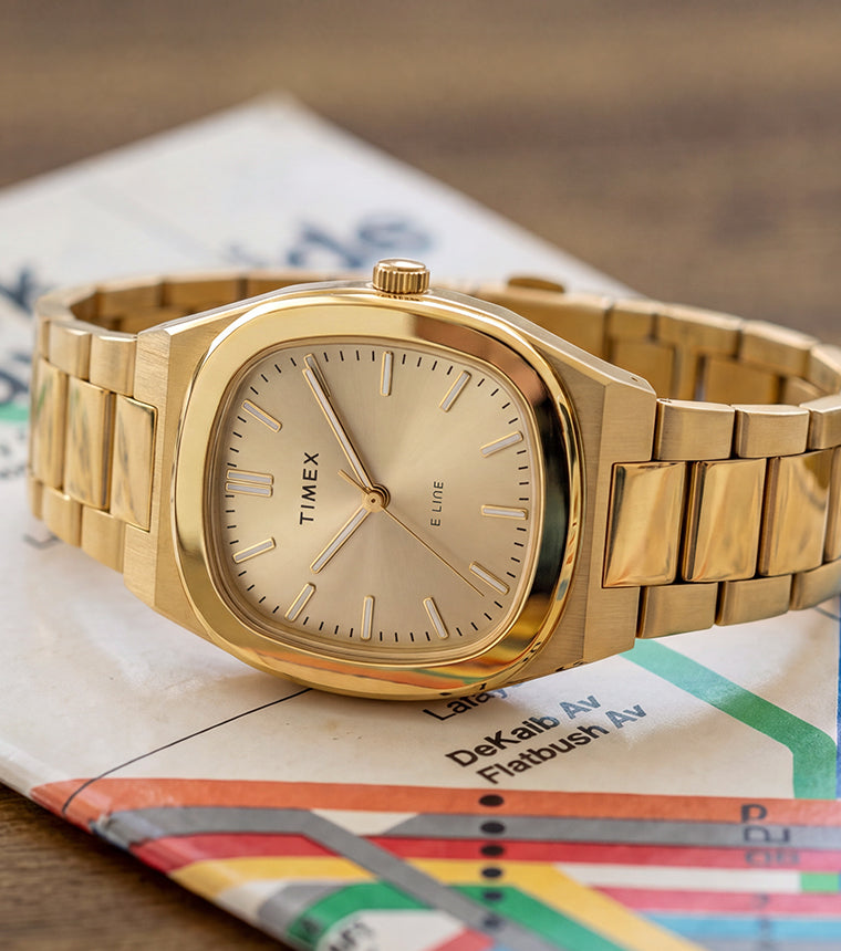 Reloj Timex® Quartz E Line de 30 mm con brazalete de acero inoxidable en tono dorado y esfera color champán, presentado sobre un mapa del metro de Nueva York.