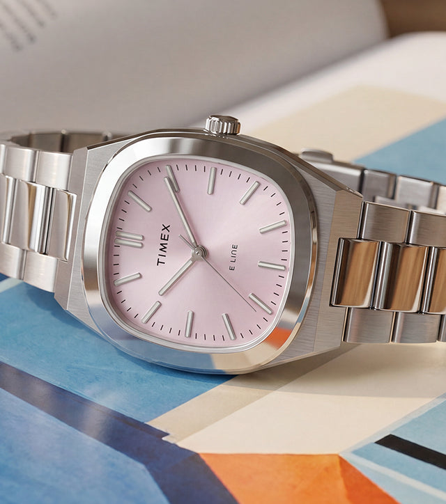 Reloj Timex® Quartz E Line de 30 mm con brazalete de acero inoxidable y esfera rosa con acabado rayos de sol, expuesto sobre un libro abierto con ilustraciones de colores.