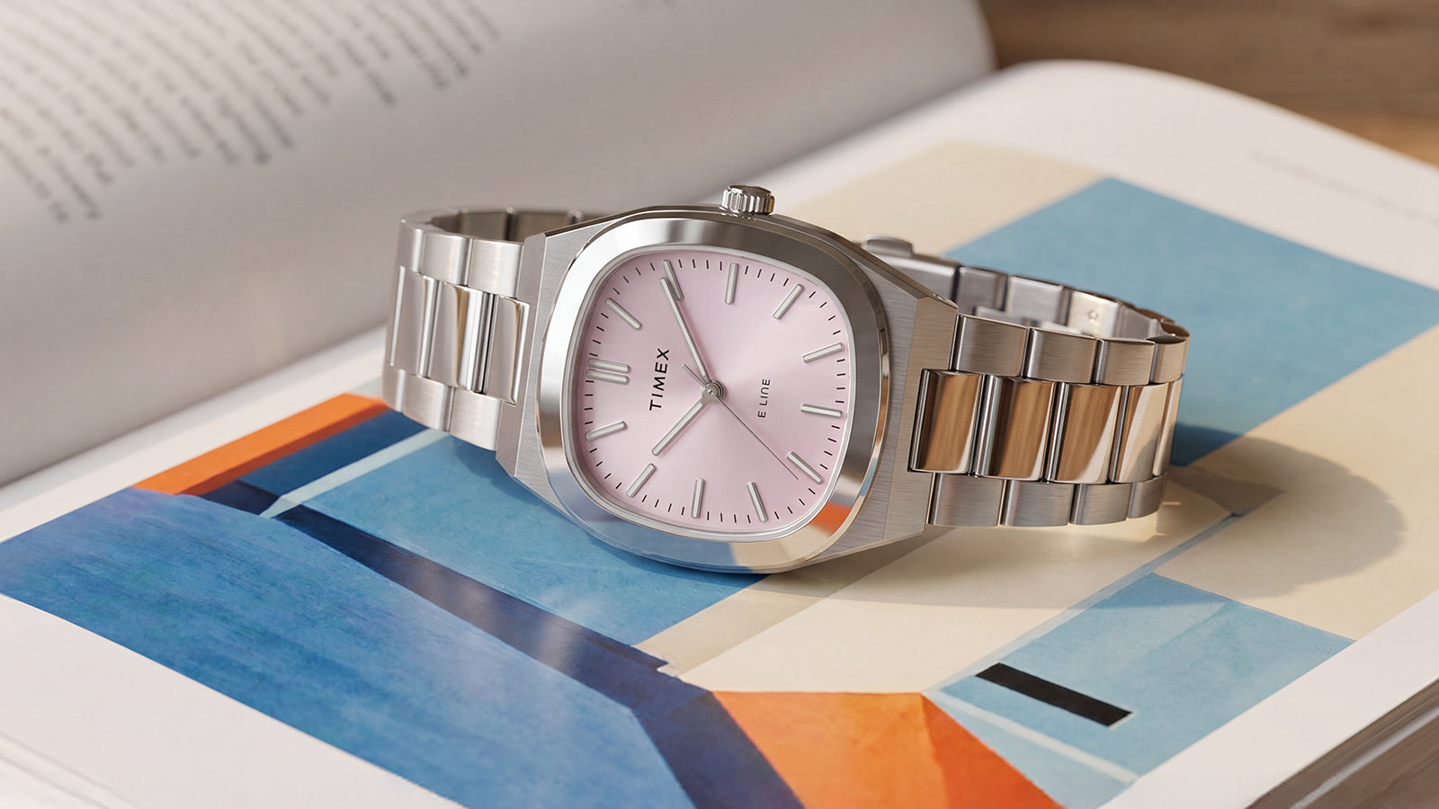 Primer plano del reloj Timex® Quartz E Line de 30 mm, de acero inoxidable y con esfera rosa, expuesto sobre un libro abierto con ilustraciones de colores.