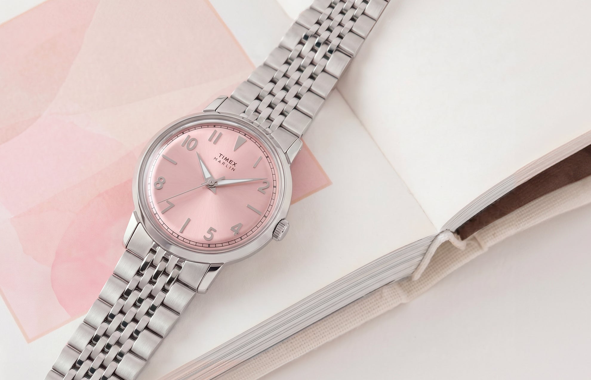 Timex Marlin Quartz watch with a pink sunray dial and stainless steel bracelet displayed on an open book with soft pink accents.