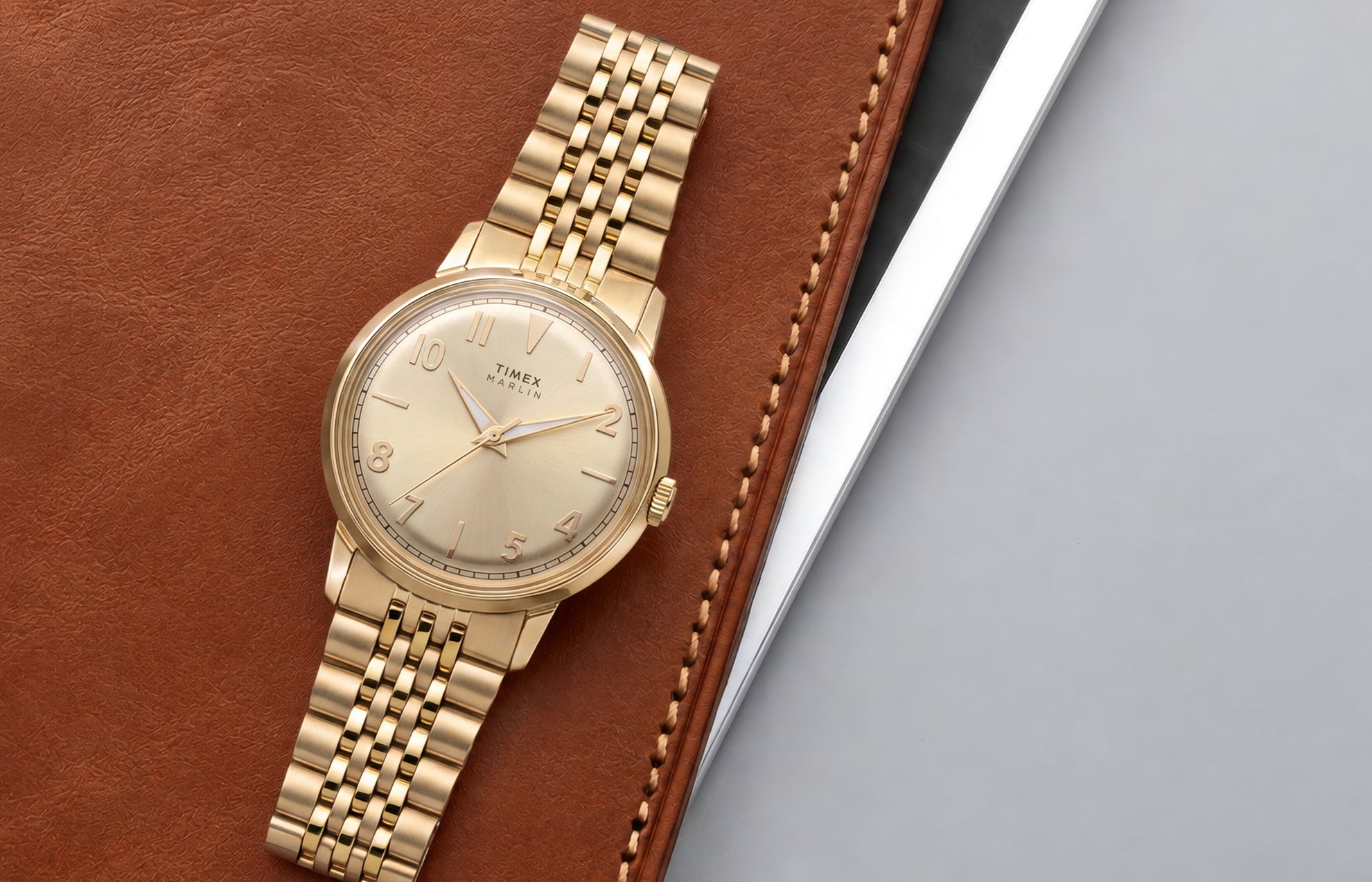 Timex Marlin Quartz watch with a champagne sunray dial and gold-tone stainless steel bracelet displayed on a leather portfolio.