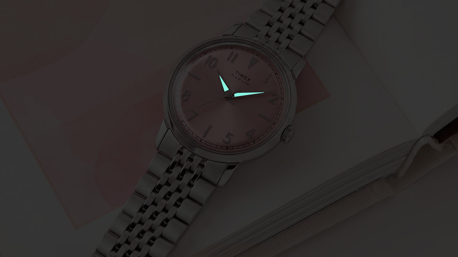 Close-up of the Timex Marlin Quartz with luminous hands illuminated on a pink sunray dial in low light.