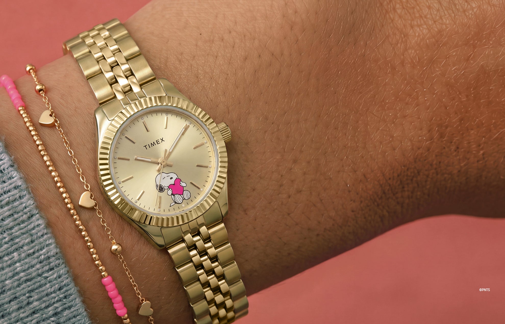 Peanuts Legacy Mini Heart Hug watch worn on the wrist, featuring a gold-tone case and bracelet, champagne dial, and Snoopy hugging a pink heart at the 6 o'clock position.