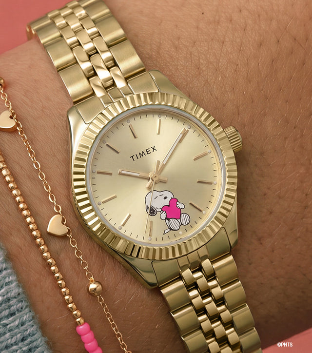 Peanuts Legacy Mini Heart Hug watch worn on the wrist, featuring a gold-tone case and bracelet, champagne dial, and Snoopy hugging a pink heart at the 6 o'clock position.
