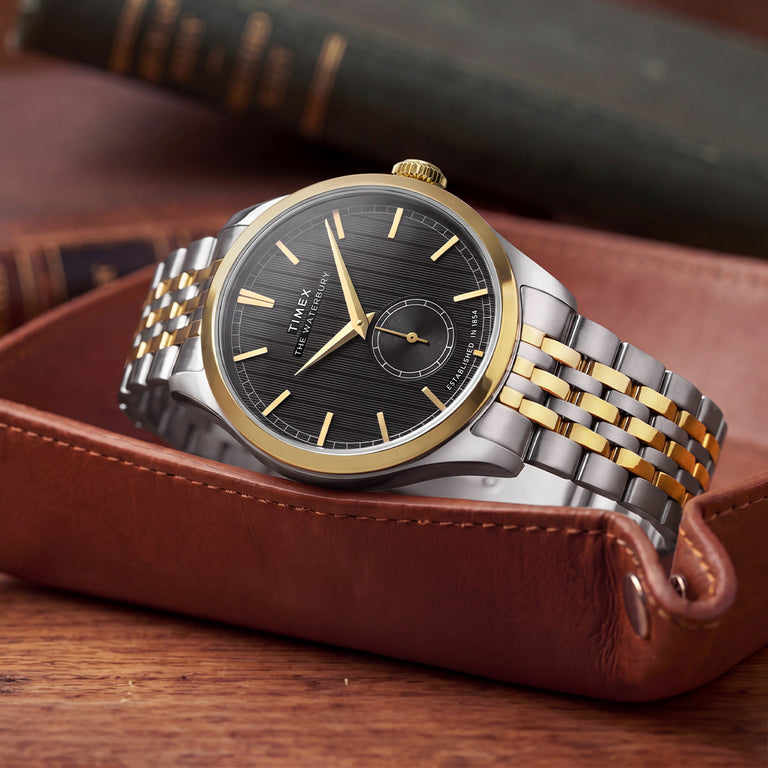 Timex Waterbury Classic watch with a two-tone stainless steel bracelet, gold-tone bezel, black textured dial, and small seconds sub-dial, displayed in a brown leather valet tray.