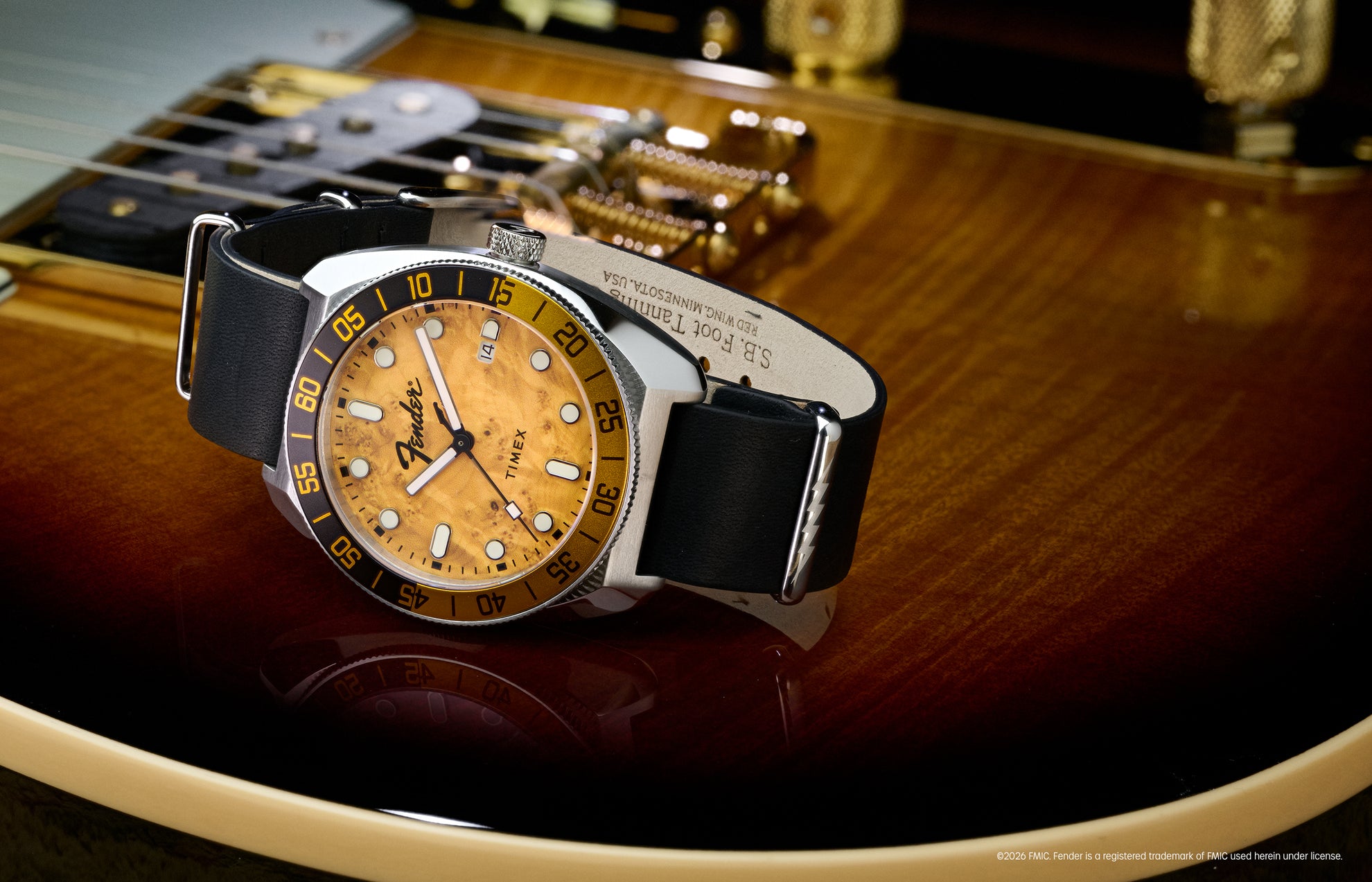 Fender x Timex Automatic watch with a silver-tone case, amber wood-grain dial, black-and-gold bezel, and black leather strap displayed on a Fender guitar.