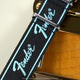 Close-up of a black Fender-branded strap with turquoise trim displayed beside an electric guitar.