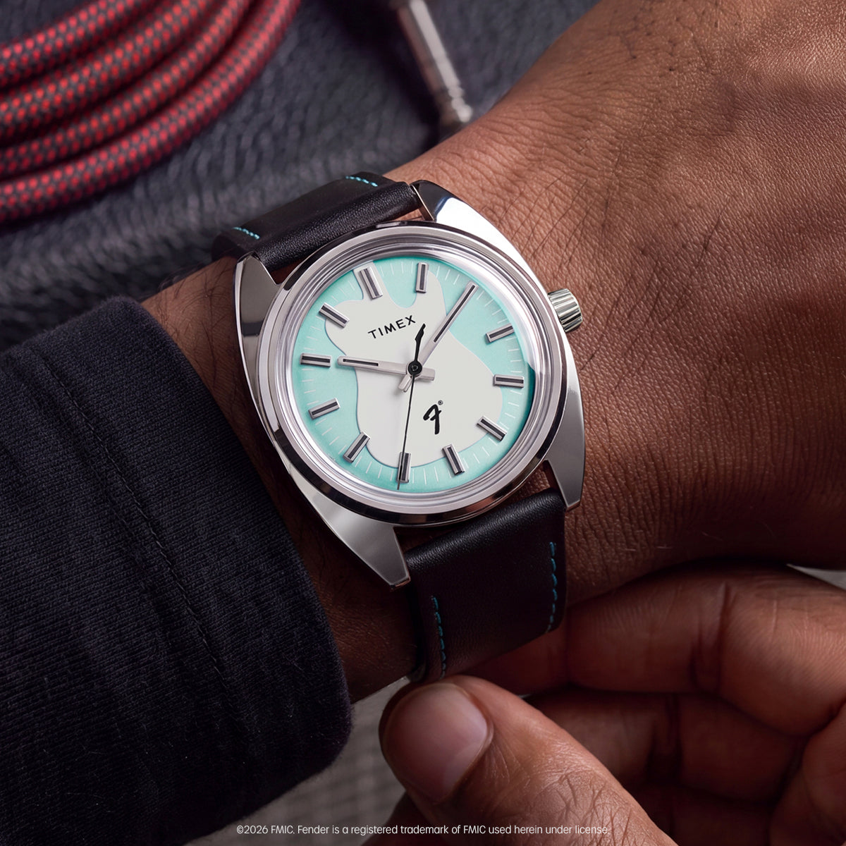 Fender x Timex watch worn on the wrist, featuring a silver-tone case, aqua dial accents, guitar motif, and black strap with turquoise stitching.