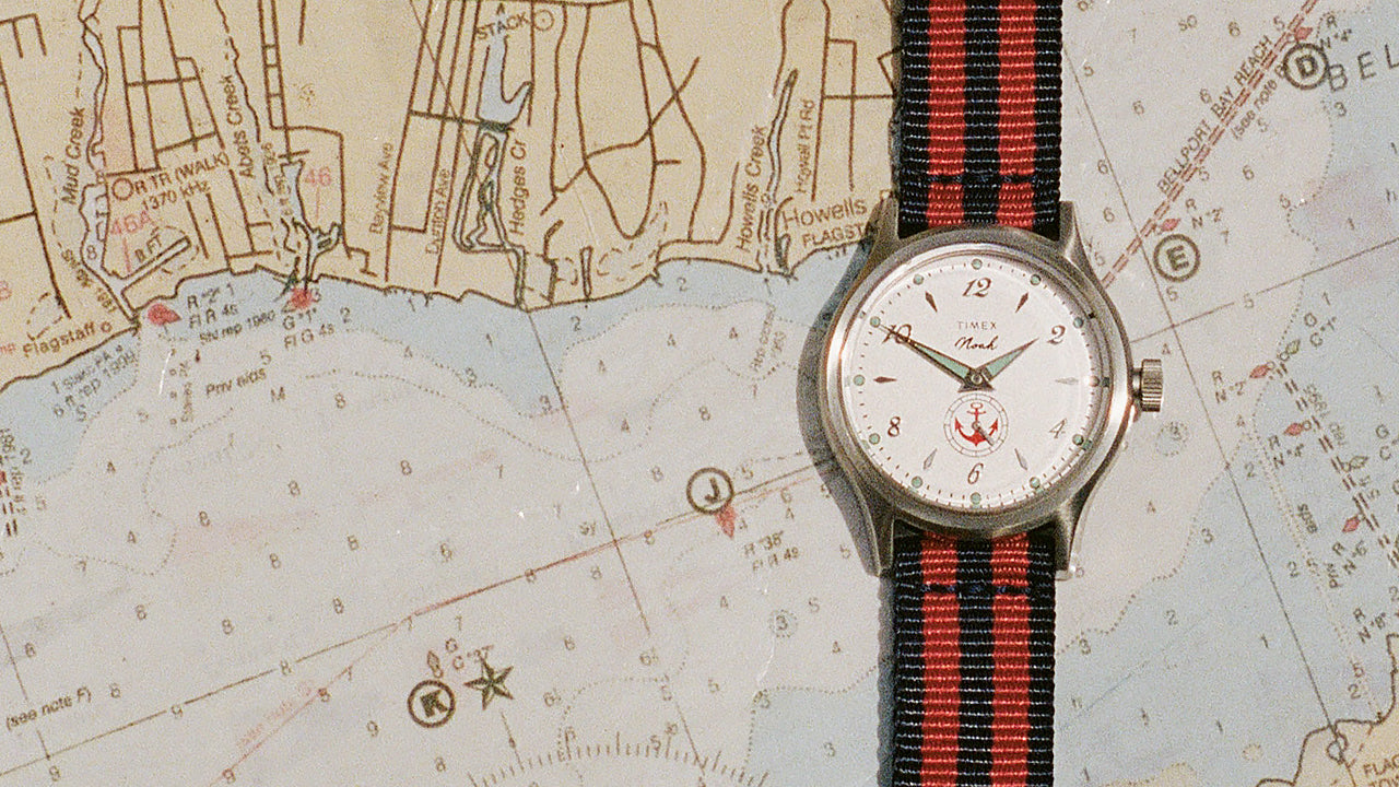 Reloj NOAH x Timex MK1 Anchor con esfera blanca y correa de tela a rayas rojas y azul marino, expuesto sobre una carta náutica.