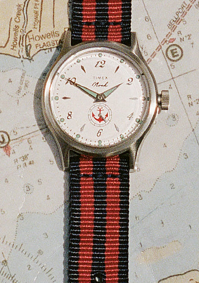 Reloj NOAH x Timex MK1 Anchor con esfera blanca y correa de tela a rayas rojas y azul marino, expuesto sobre una carta náutica.