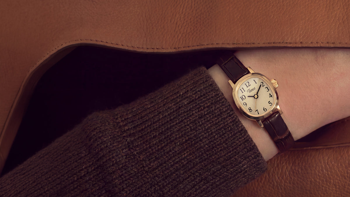 Reloj Cavatina® Mini lucido en la muñeca, bajo una manga de punto marrón y un abrigo de color beige.