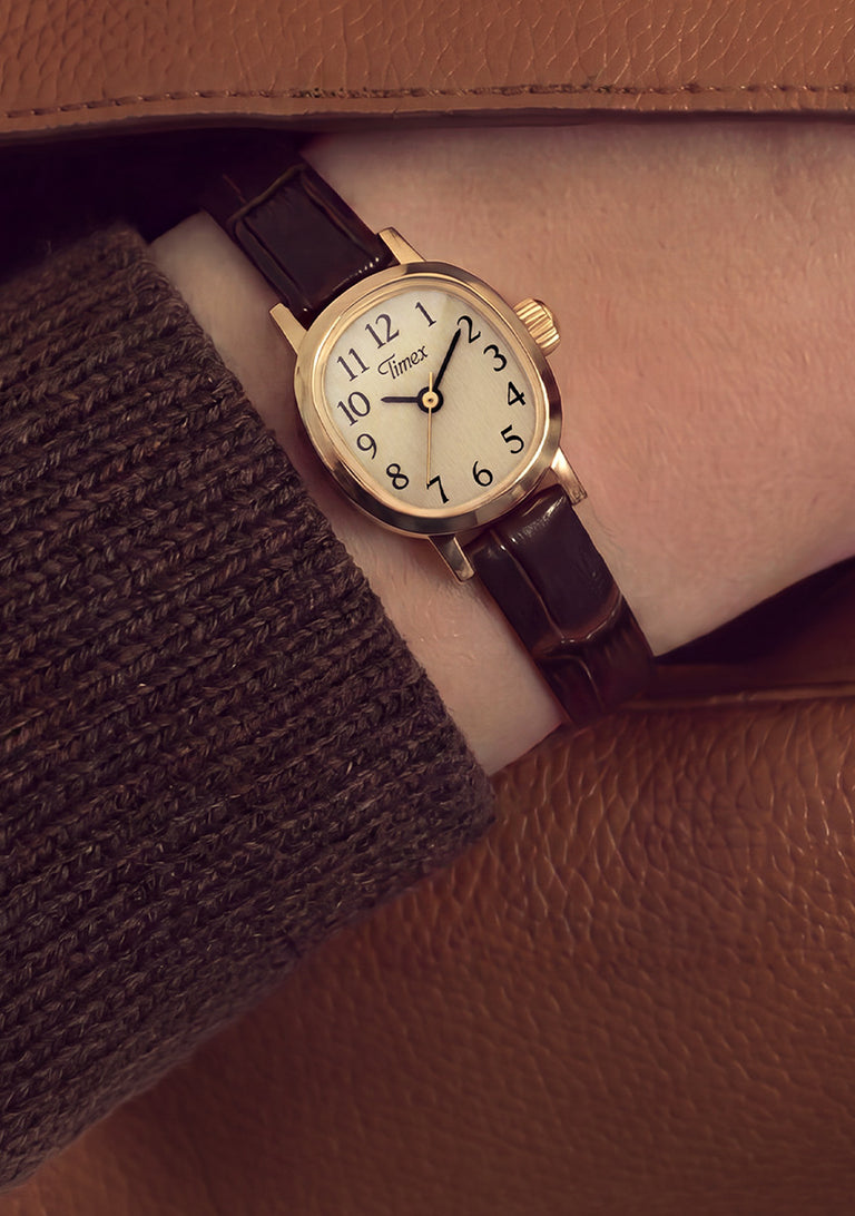 Reloj Cavatina® Mini lucido en la muñeca, bajo una manga de punto marrón y un abrigo de color beige.