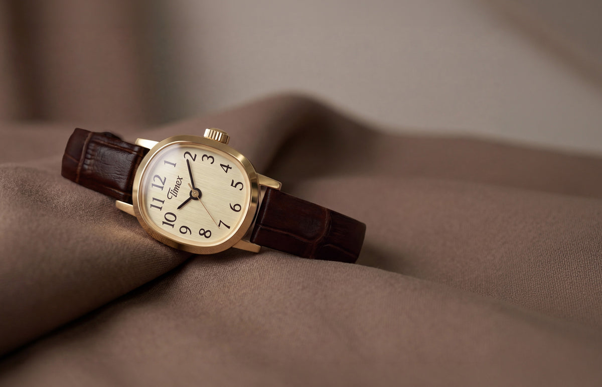 Reloj Cavatina® Mini con caja en tono dorado, esfera color champán y correa de piel marrón oscuro sobre un suave tejido marrón.