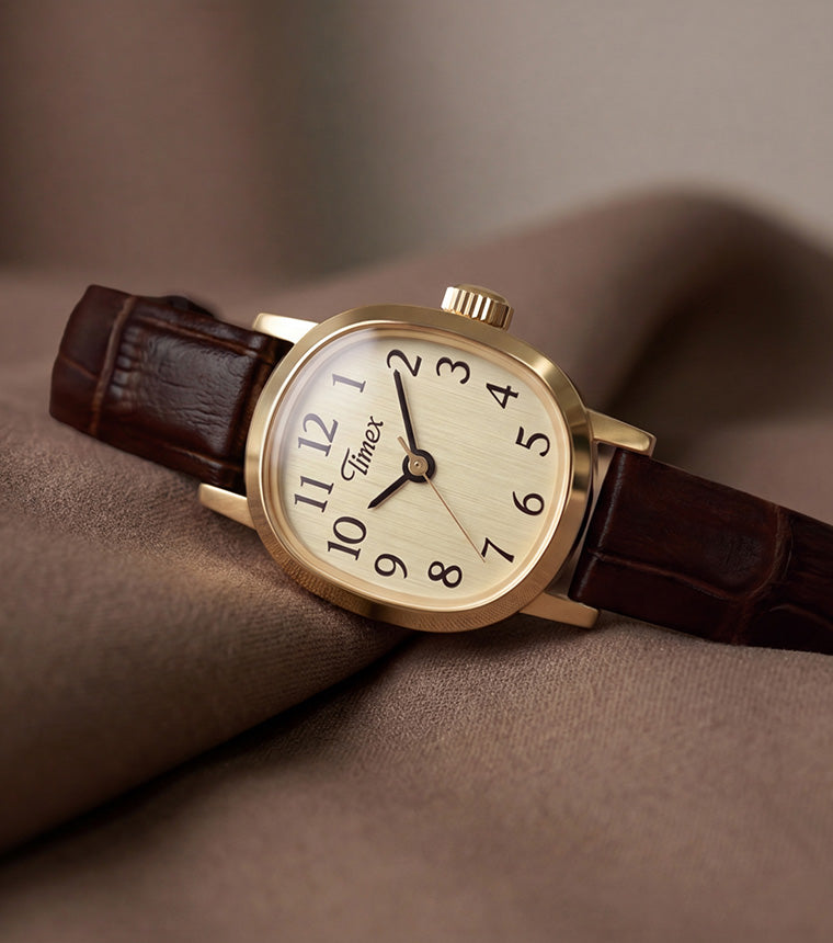 Reloj Cavatina® Mini con caja en tono dorado, esfera color champán y correa de piel marrón oscuro sobre un suave tejido marrón.