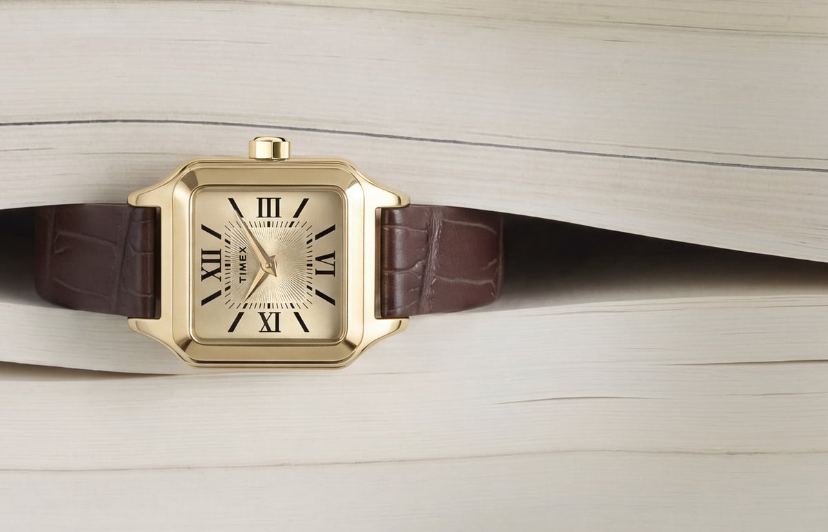 Timex Addison watch with a gold-tone rectangular case, champagne dial, black Roman numerals, and brown leather strap displayed on an open book.
