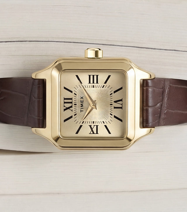Timex Addison watch with a gold-tone rectangular case, champagne dial, black Roman numerals, and brown leather strap displayed on an open book.