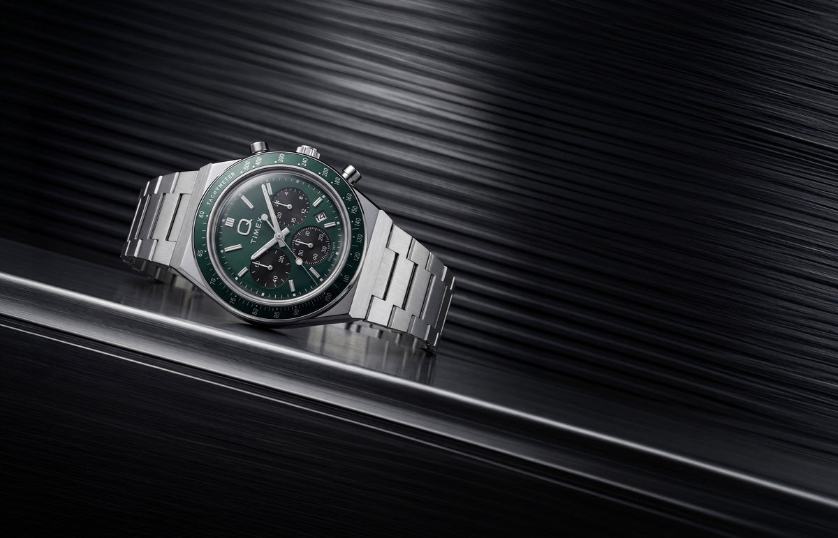 Q Timex Chronograph with a green dial and stainless-steel bracelet displayed on a dark metallic surface.