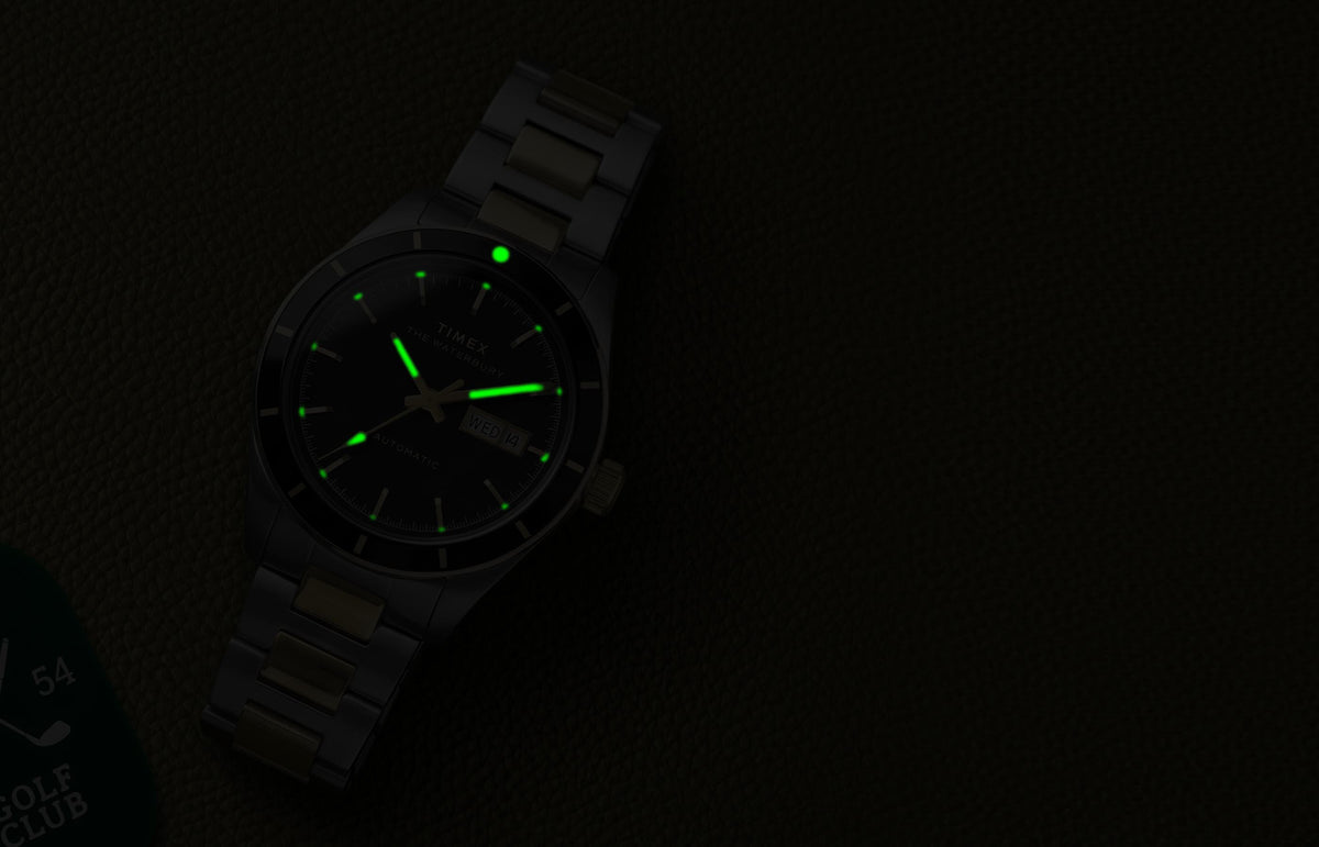 Waterbury Heritage Automatic watch illuminated by green glowing hands and hour markers on textured leather in low light.
