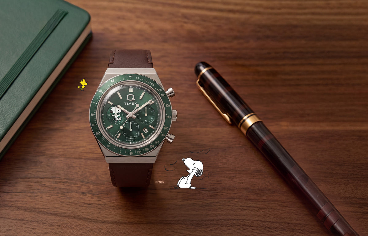 Cronógrafo Peanuts® x Q Timex con esfera verde, subesfera de Snoopy y correa de piel marrón, expuesto junto a un cuaderno y un bolígrafo.