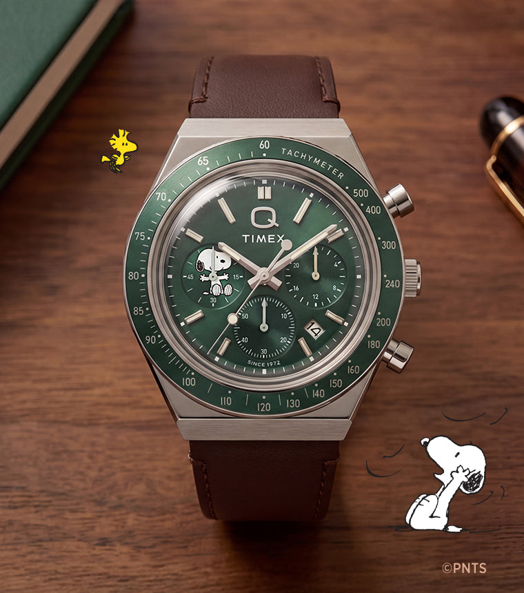 Cronógrafo Peanuts® x Q Timex con esfera verde, subesfera de Snoopy y correa de piel marrón, expuesto junto a un cuaderno y un bolígrafo.